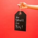 Close-up of hand holding a 'Shop Small Help Big' tag against a red background, promoting small businesses.